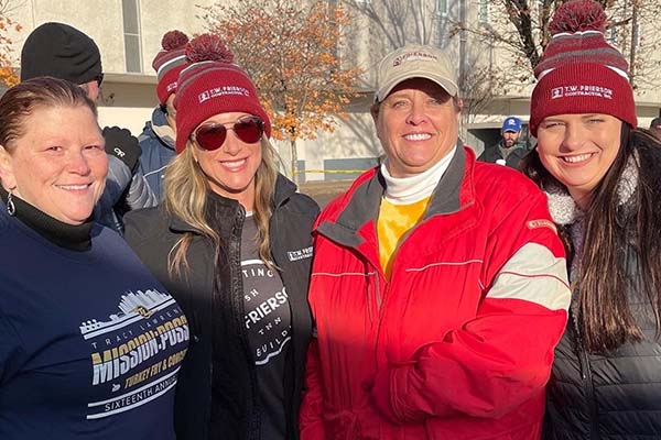 Four women in winter hats and jackets smile together at an outdoor community event, standing in front of other attendees on a sunny day.